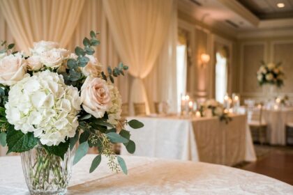 Composition florale élégante pour décoration de salle de mariage avec roses roses pâles, pivoines blanches et feuillage d'eucalyptus dans un vase cristal sur nappe damassée