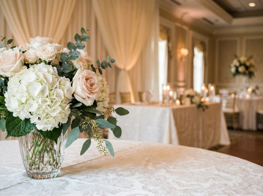Composition florale élégante pour décoration de salle de mariage avec roses roses pâles, pivoines blanches et feuillage d'eucalyptus dans un vase cristal sur nappe damassée