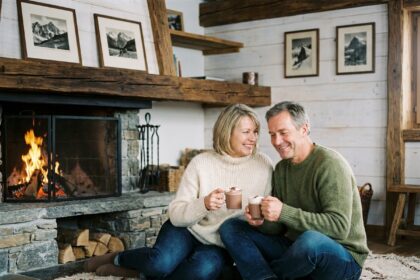 Couple en pull d'hiver assis près d'une cheminée en pierre dans un chalet montagnard chaleureux, tenant des tasses de chocolat chaud